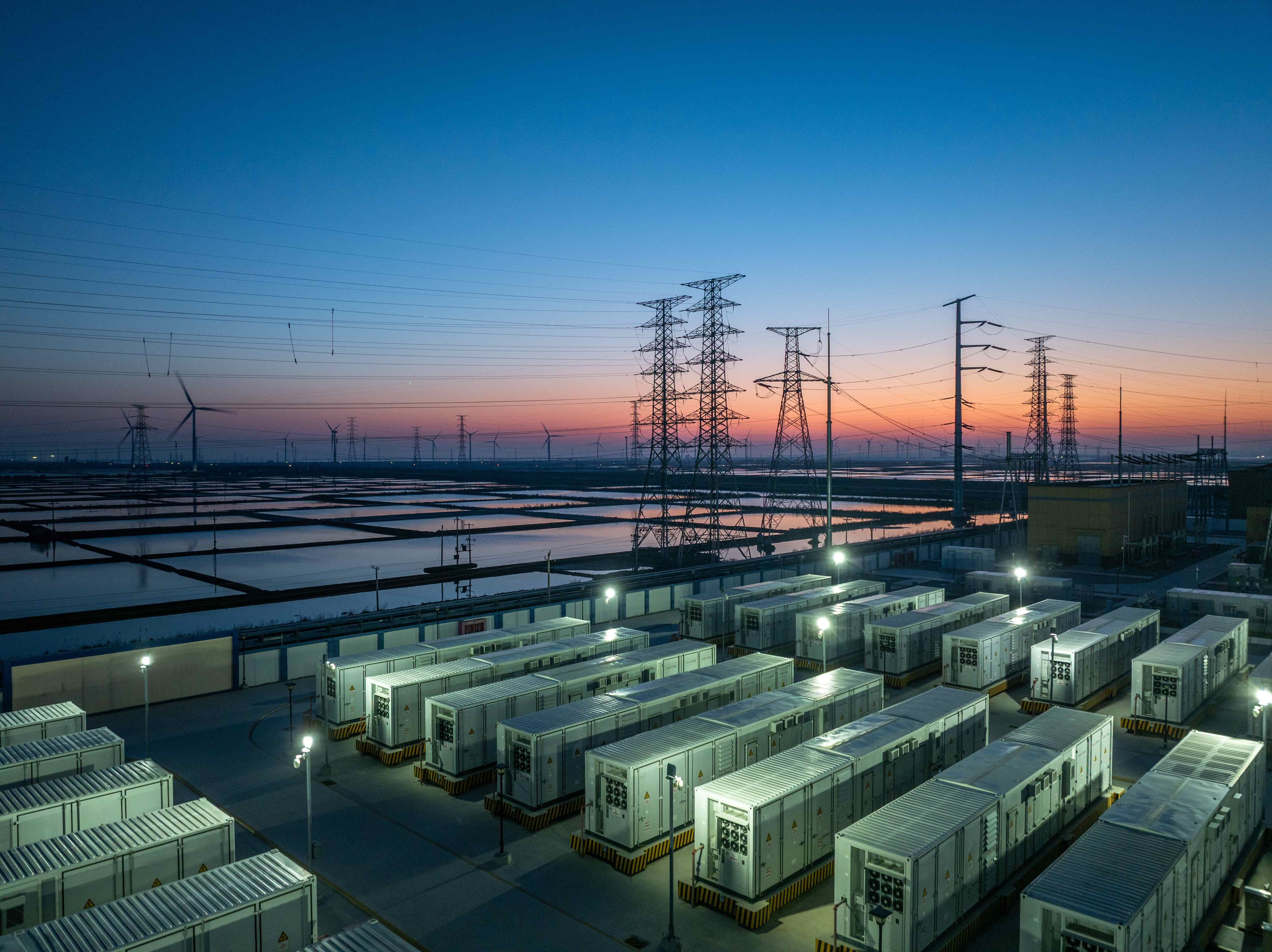 Battery energy storage site beneath high-voltage pylons and wind turbines at dusk